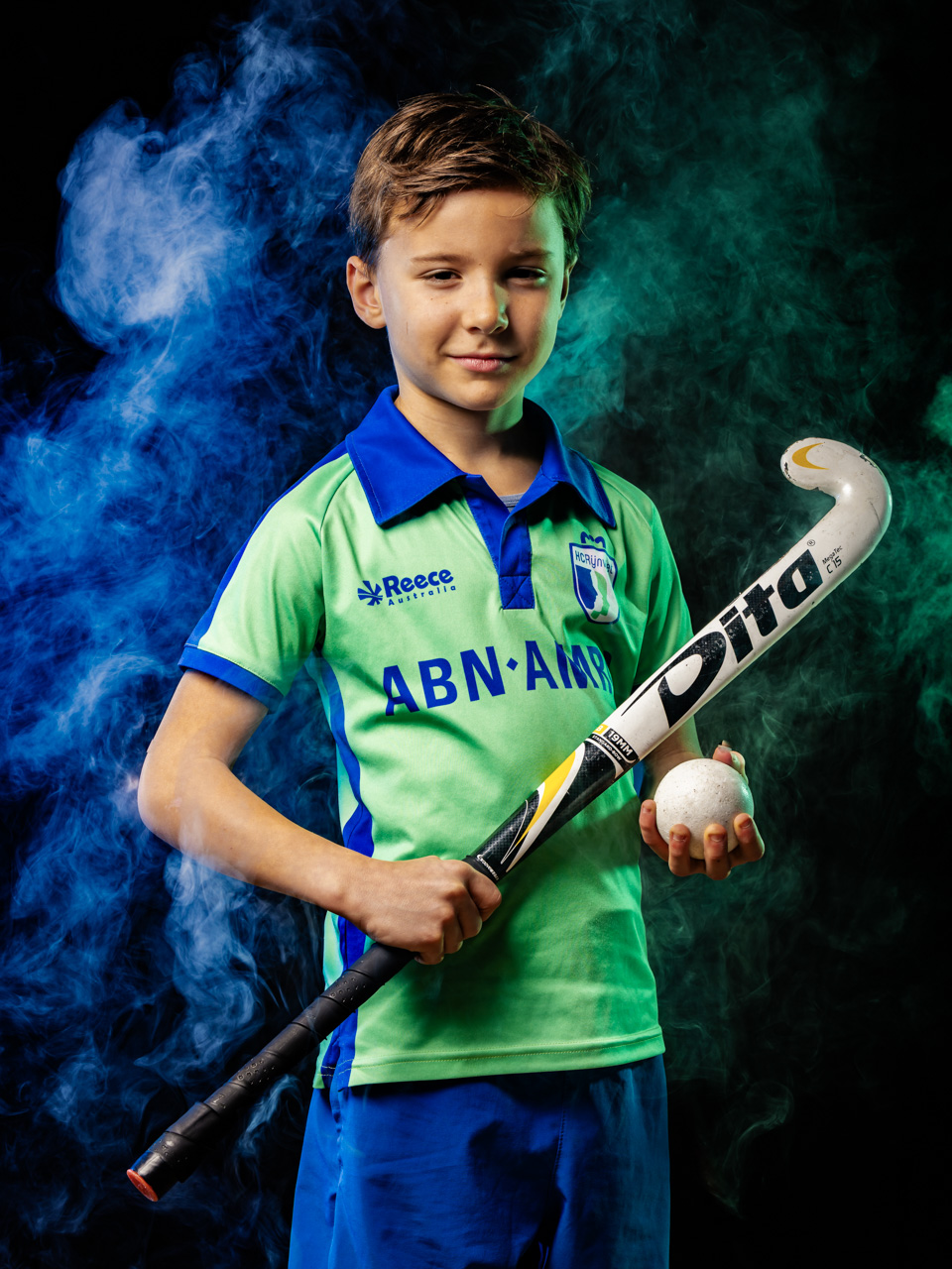 Dynamisch PhiPho Hockey Portret HC Rijnvliet met off-camera flash. Jongen draagt het groen-blauwe clubtenue, gesponsord door ABN AMRO en heeft een Dita hockeystick en bal vast. Gefocuste blik en is geportretteerd tegen een donkere studioachtergrond. Blauw en Groen rook versterkt het portret.