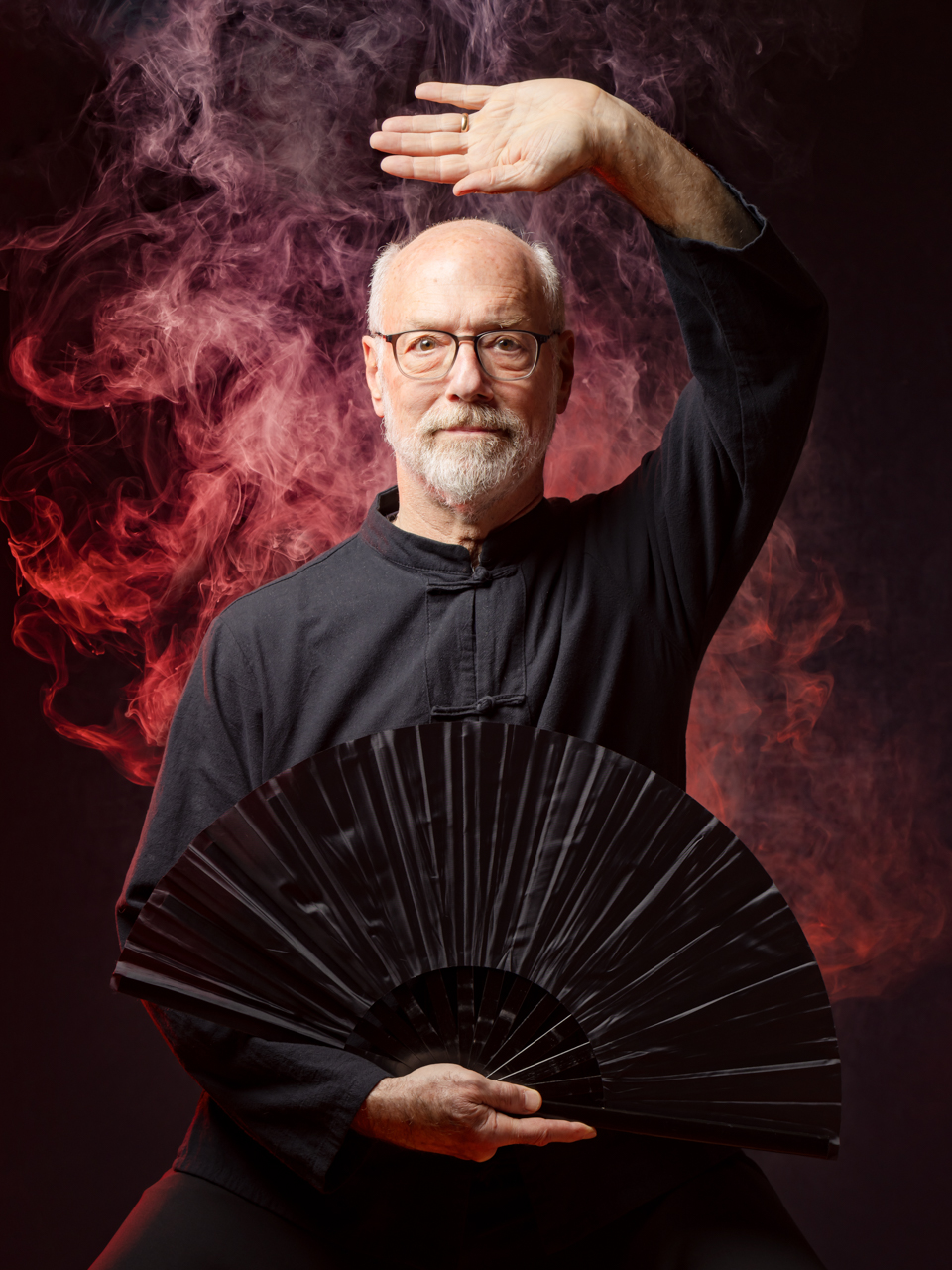 Dynamisch PhiPho Tai Chi Portret van Mitchell Sandler van Taiji Tolsteeg. Mitchell draagt zwarte tai chi kleding heeft een zwarte waaier vast. Gefocuste blik en is geportretteerd tegen een donkere studioachtergrond. Rode rook versterkt het portret en symboliseert vuur.