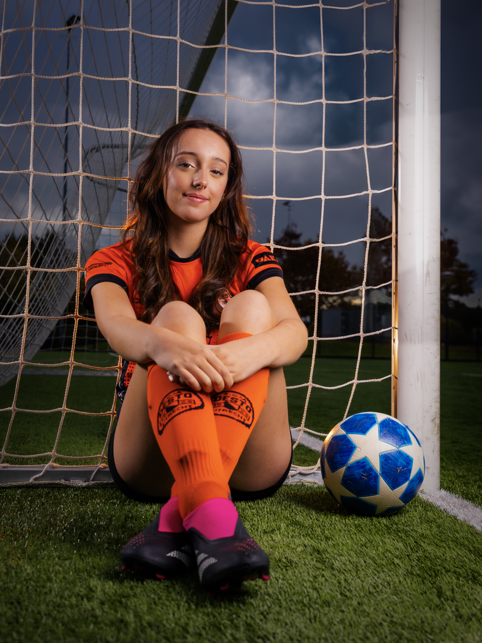Dynamisch PhiPho Voetbal Portret van Zoë C.O.V. Desto met off-camera flash. Zoe zit naast het doel en draagt het knaloranje-zwart clubtenue en sokken, van sportmerk Craft en DAKA, sponsor Ronald McDonald Huis Utrecht, adidas voetbalschoenen en heeft een Champions League bal. Zoe kijkt met een zachte blik en is geportretteerd tegen een dramatische donkere lucht door gebruik van flitslicht overdag.