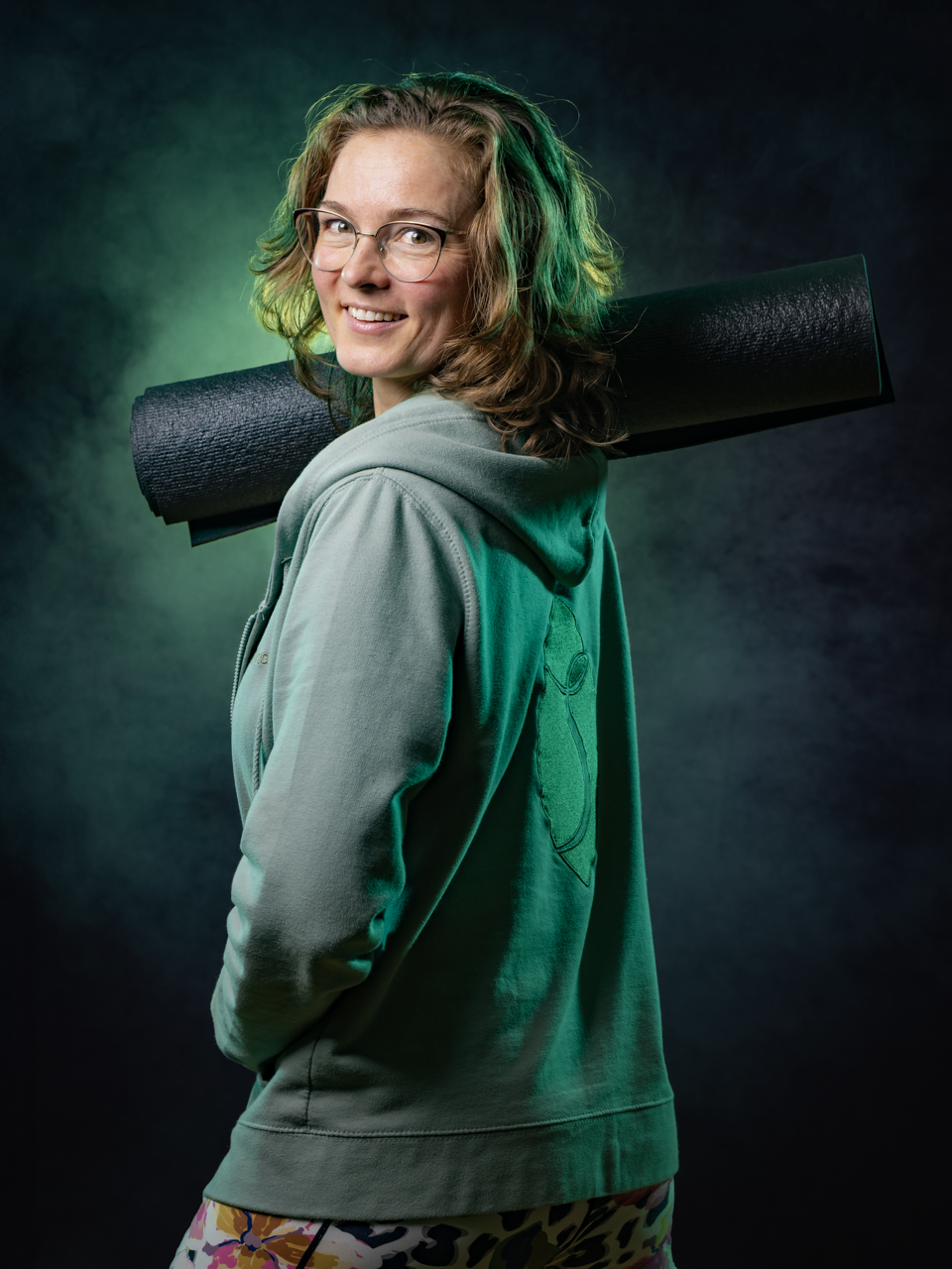 Dynamisch PhiPho Yoga Portret van Karianne Hol van YogaShape Utrecht in Hoograven / Lunetten. Karianne draagt groene YogaShape kleding en heeft een zwarte yogamat vast. Blik achteruit over de schouder en is geportretteerd tegen een donkere studioachtergrond. Groene nevel versterkt het portret.